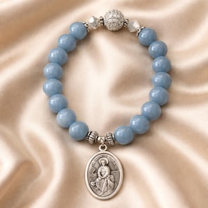 May include: A light blue beaded bracelet with a silver pendant. The bracelet features large, round beads and a decorative silver clasp. The pendant has an oval shape with an image of a saint. The bracelet is displayed on a satin surface.