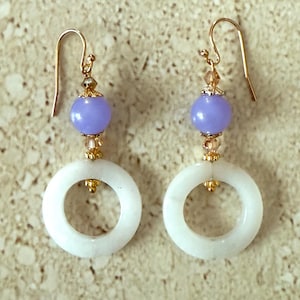 May include: A pair of dangle earrings featuring white circular pendants and lavender beads. The earrings have gold-colored hooks and accents, with small crystal-like beads. The earrings are designed for pierced ears.