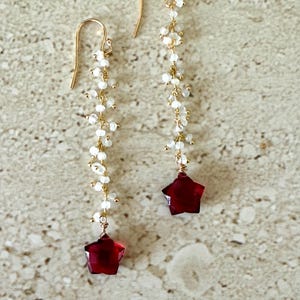 May include: A pair of long, dangling earrings with a gold chain and white beads. The earrings have a red star-shaped charm at the bottom.