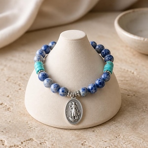 May include: A beaded bracelet featuring blue and white speckled beads, turquoise cylinder beads, and a silver pendant. The pendant depicts a religious figure. The bracelet is displayed on a cream-colored stand.