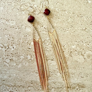 May include: A pair of gold-colored dangle earrings. Each earring features a square, dark red gemstone at the top, connected to a geometric gold frame. Long, delicate gold chains cascade from the frame, creating a flowing, elegant design.