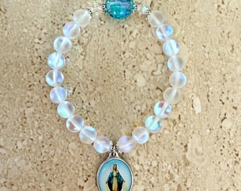 May include: A white iridescent beaded bracelet with a blue crystal charm and a silver charm featuring an image of the Virgin Mary.