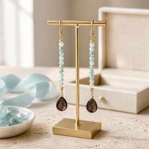 May include: A pair of gold-colored dangle earrings. Each earring features a strand of light blue beads, a gold accent, and a teardrop-shaped, dark brown gemstone. The earrings are displayed on a gold-colored stand.