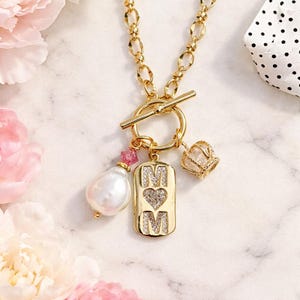 May include: Gold-toned necklace with a toggle clasp, featuring a pendant with the word "MOM" and a heart, a pearl, and a crown charm. The necklace has a chain link design and is adorned with pink accents.