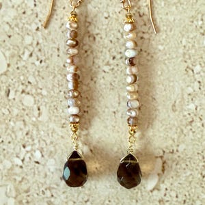 May include: A pair of dangle earrings featuring a strand of small, light-colored beads with gold-colored accents. Each earring has a dark brown, teardrop-shaped gemstone pendant and gold-colored ear wires.