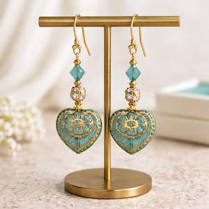 May include: A pair of heart-shaped earrings displayed on a gold stand. The earrings feature a teal and gold floral design, with a clear blue crystal and a gold-colored rhinestone ball. The earrings are suspended from gold-colored hooks.