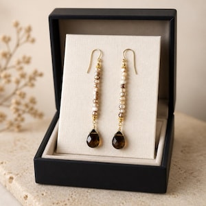 May include: A pair of gold-toned earrings displayed in a black jewelry box. Each earring features a strand of small, off-white and brown beads, with a teardrop-shaped, dark brown gemstone at the bottom. The earrings are presented against a cream-colored backdrop.