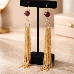 May include: A pair of gold-toned earrings with a red gemstone and cascading chain fringe. The earrings are displayed on a black stand, showcasing their intricate design and elegant style. The gemstones are square-shaped.