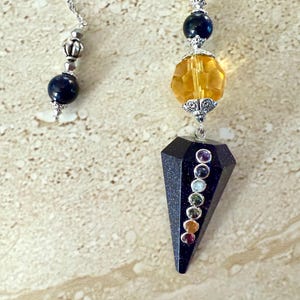 May include: Two pendulums with silver chains. One features a dark blue bead and silver accents. The other has a faceted yellow bead, a dark blue bead, and a black, faceted, teardrop-shaped stone with small, colorful gemstones.
