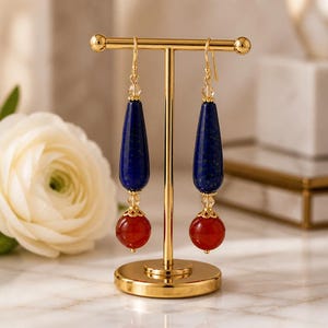 May include: A pair of drop earrings hanging on a gold-toned T-bar stand. Each earring features a long, teardrop-shaped dark blue stone topped with a small clear bead, followed by a round, smooth red stone at the base with gold-toned metal accents.