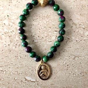 May include: A green and pink gemstone bracelet with a gold charm featuring a religious figure. The bracelet is made of round beads and has a gold clasp.