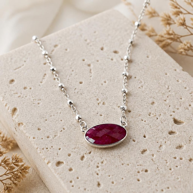 May include: A silver chain necklace featuring a faceted oval-shaped deep red gemstone pendant. The chain is composed of small, evenly spaced metallic beads integrated into the links, connecting to the bezel-set stone at the center of the piece.