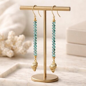 May include: Gold-tone earrings with a dangling design. Each earring features a strand of light blue beads, a gold accent, and a gold seashell charm. The earrings are displayed on a gold stand.