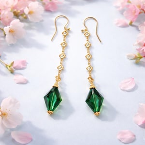 May include: A pair of gold-colored dangle earrings. Each earring features a series of small, white flower-shaped accents connected by a delicate chain. The earrings terminate with a faceted, emerald-green, diamond-shaped crystal. Pink flowers and petals are in the background.