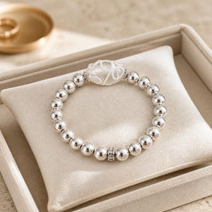 May include: A silver beaded bracelet with a clear, carved centerpiece and small, sparkling accents. The bracelet is displayed on a beige pillow inside a jewelry box. The bracelet is a stylish accessory.