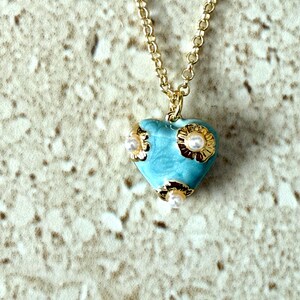 May include: A gold-colored chain necklace with a pendant. The pendant is a light blue heart shape with gold-colored accents and small white pearls. The necklace is displayed against a textured, light-colored background.