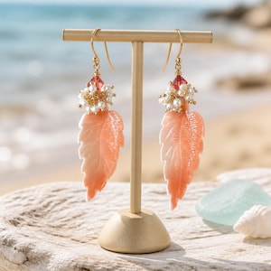May include: A pair of dangle earrings featuring carved peach-colored leaf-shaped pendants, accented with clusters of small white pearls and a single red bead at the top, suspended from gold-toned metal hooks on a T-shaped display stand.