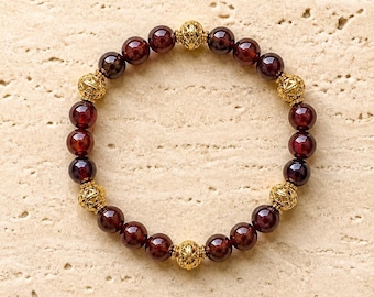 May include: A circular beaded bracelet featuring smooth, deep red garnet gemstone beads alternating with ornate, spherical gold-toned metal spacers. The beads are arranged in a repeating pattern, creating a symmetrical design with a polished finish.