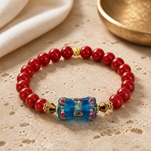 May include: Red beaded bracelet with a central blue glass bead. The bracelet features small red beads and gold-colored accents. The blue bead has red and green details. The bracelet is displayed on a light-colored surface.