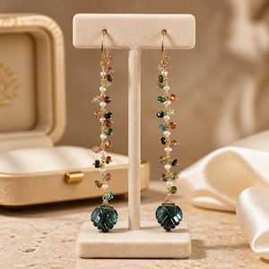 May include: Gold-tone dangle earrings with a cascade of colorful gemstones and small pearls. Each earring features a faceted, dark green, leaf-shaped stone at the bottom. The earrings are displayed on a beige stand.