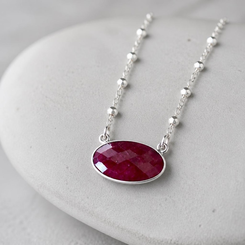 May include: A silver chain necklace featuring a faceted, oval-shaped deep red gemstone pendant. The chain is designed with small, evenly spaced silver beads along its length, connecting to the central ruby-colored stone set in a silver frame.