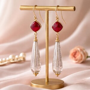 May include: A pair of gold-toned earrings with red square-cut gemstones and long, silver teardrop-shaped pendants. The earrings are displayed on a gold stand against a soft pink background, with a pearl necklace and a rose in the background.