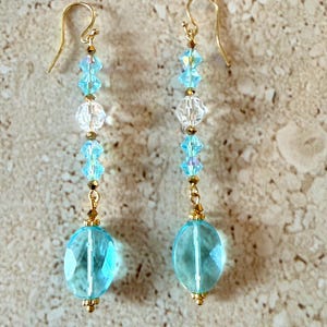 May include: A pair of dangle earrings with gold-colored hooks. Each earring features a series of light blue and clear crystal beads, culminating in a large, oval, light blue faceted bead. The earrings are designed for pierced ears.