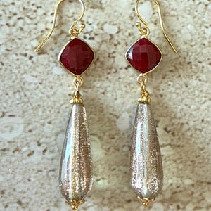 May include: A pair of gold-colored earrings. Each earring features a square, faceted, deep red gemstone at the top, connected to a teardrop-shaped, silver-colored bead with gold glitter accents. The earrings have a hook closure.