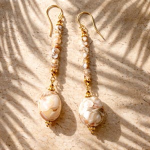May include: A pair of dangle earrings with gold-colored hooks and accents. Each earring features a strand of small, light-colored beads leading to a larger, oval-shaped pendant with a cream and brown pattern. The earrings are displayed on a textured, light-brown surface.