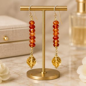 May include: Gold-toned dangle earrings with a vertical row of orange-red beads and a faceted yellow-gold sphere at the bottom. The earrings are displayed on a gold stand, showcasing their design and color.