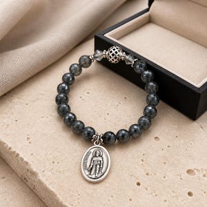 May include: A beaded bracelet featuring dark gray, faceted beads. A silver-toned oval charm with an image of a saint is attached. The bracelet is displayed near a black jewelry box with a beige interior.
