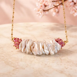 May include: A gold chain necklace featuring a central bar of white, irregular-shaped pearls. The necklace has clusters of pink gemstones on each end. The background is a soft pink with a hint of a floral branch.