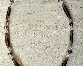May include: A necklace featuring alternating dark brown, elongated beads and clear, faceted crystals. The necklace is strung on a silver-toned chain and has a silver-toned clasp. The beads are arranged in a symmetrical pattern.