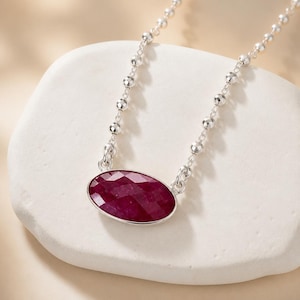 May include: A silver necklace featuring a faceted, oval-shaped deep red gemstone pendant. The chain is composed of small, round, reflective silver beads that connect to the central setting holding the polished, dark crimson stone securely.