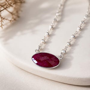 May include: A necklace featuring a faceted, oval-shaped deep red ruby gemstone set in a silver bezel. The pendant is attached to a delicate silver chain composed of small, round, reflective metallic beads linked together in a continuous pattern.