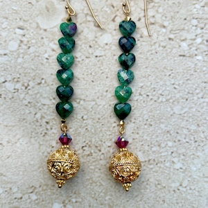 May include: A pair of long, dangling earrings with a gold chain and green heart-shaped beads. The earrings have a gold ball at the bottom with a small, clear crystal bead above it.