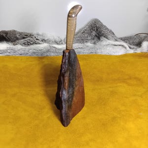 以下が含まれることがあります： 木製の柄と、暗く質感のある木製の台座が付いた手作りのナイフ。柄は薄茶色で、蛇の頭に似たカーブを描いています。台座は暗く、粗く削られた木片です。ナイフは黄色の表面に展示されています。