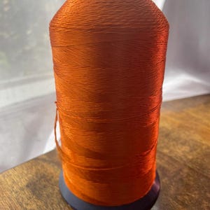 May include: A spool of bright orange thread. The thread is tightly wound around a cylindrical core with a black top and a dark blue base. The thread has a slight sheen and appears smooth.