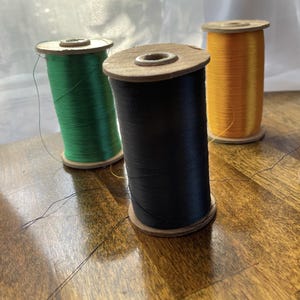 May include: Three spools of thread in green, black, and orange. The thread is wound around wooden spools. The spools are arranged on a wooden surface.