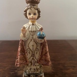 May include: A religious figurine of a child wearing a crown, a white robe with gold-colored embroidery, and a dusty rose cape. The figure holds a small blue orb topped with a cross in one hand and raises the other hand in a gesture of blessing.