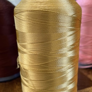 May include: A spool of golden-yellow thread, tightly wound, with a dark blue base. The thread has a slight sheen, suggesting a silk or satin material. Other spools of thread in shades of brown and pink are visible in the background.