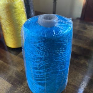 May include: A close-up of three spools of thread. The front spool is bright blue, wrapped in clear plastic. Behind it are spools of yellow and black thread. The spools are cylindrical with a gray center.