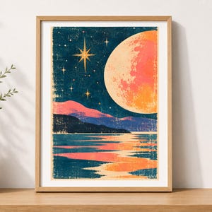 May include: A framed wall art print featuring a dark blue night sky with a large orange moon, bright stars, and pink-tinted mountains reflecting over a calm body of water. The artwork has a textured, distressed finish within a light wood frame.