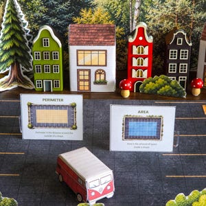 Puede incluir: Un diorama con casas de papel en verde, blanco, rojo y marrón, y una furgoneta roja y blanca. Las tarjetas muestran "PERIMETER" y "AREA" con definiciones. La escena está ambientada sobre un fondo de árboles y una carretera.