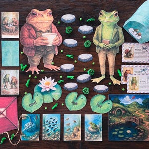 May include: A whimsical collection of illustrated cards and cutouts featuring anthropomorphic frogs, lily pads, and a charming landscape. Includes a red kite, a teal container, and cards with the words "snail", "toad", and "frog".