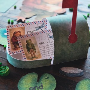 May include: A green mailbox with a red flag contains illustrated cards with frogs and dragonflies. The cards are labelled "TOAD" and "FROG". Additional game pieces include lily pads and a stone with the word "lonely".