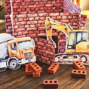 Op de afbeelding: Een speelgoed-bouwplaats met een gele graafmachine, een cementwagen en rode bakstenen blokken. De graafmachine graaft in een bakstenen muur, en de cementwagen staat ernaast. De scène speelt zich af op een houten oppervlak.
