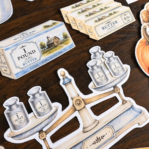 May include: A collection of die-cut stickers featuring a vintage balance scale with one-ounce weights, sticks of butter labeled 4oz and 1 pound, a thermometer, and a pumpkin. The illustrations use a watercolor style with blue and cream tones.