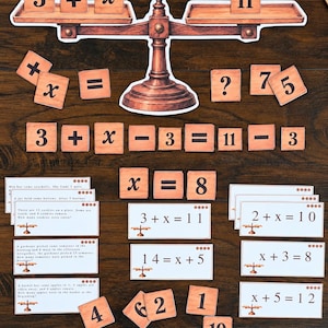 May include: A wooden balance scale cutout with wooden tiles displaying numbers and mathematical symbols including 3, +, x, 11, =, 8, 7, 5, 4, 2, 1, 10, 14, and 12. Several white rectangular cards feature printed word problems and algebraic equations.