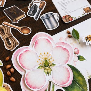May include: A collection of educational paper cutouts featuring a large pink and white apple blossom, a Holy Bible, a flint and steel, a metal cup, a wooden spoon, a coil of rope, and a Saddlebag Challenge card with text and illustrations.